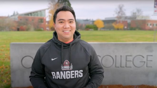 Employee in gray OC Rangers sweatshirt standing in front of concrete college sign.