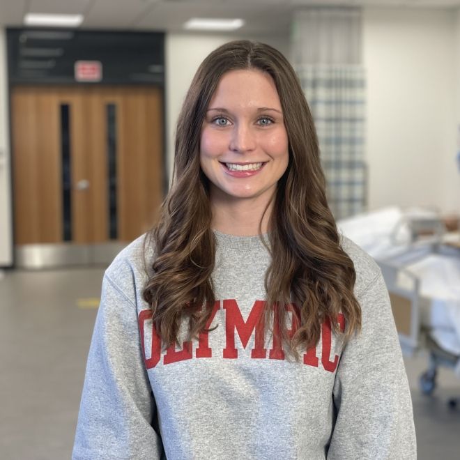Student in gray "Olympic" crewneck smiling in nursing simulation lab.