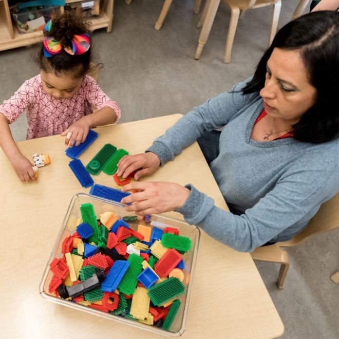 Looking down at a table with child blocks. Teacher is sitting and playing with blocks with a little girl.