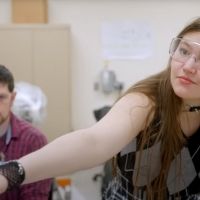 Student wearing lab goggles, passing a lego. Person in background working at computer.