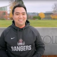 Employee in gray OC Rangers sweatshirt standing in front of concrete college sign.