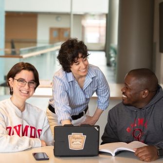 A faculty member with two students on either side of them, all looking at a tablet computer on the table in front of them.