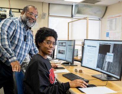 Student and instructor smiling at camera as they work at a computer station