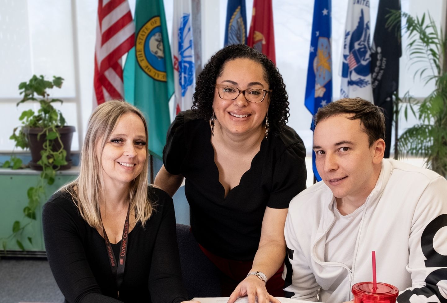 Three people at table with flags in background.