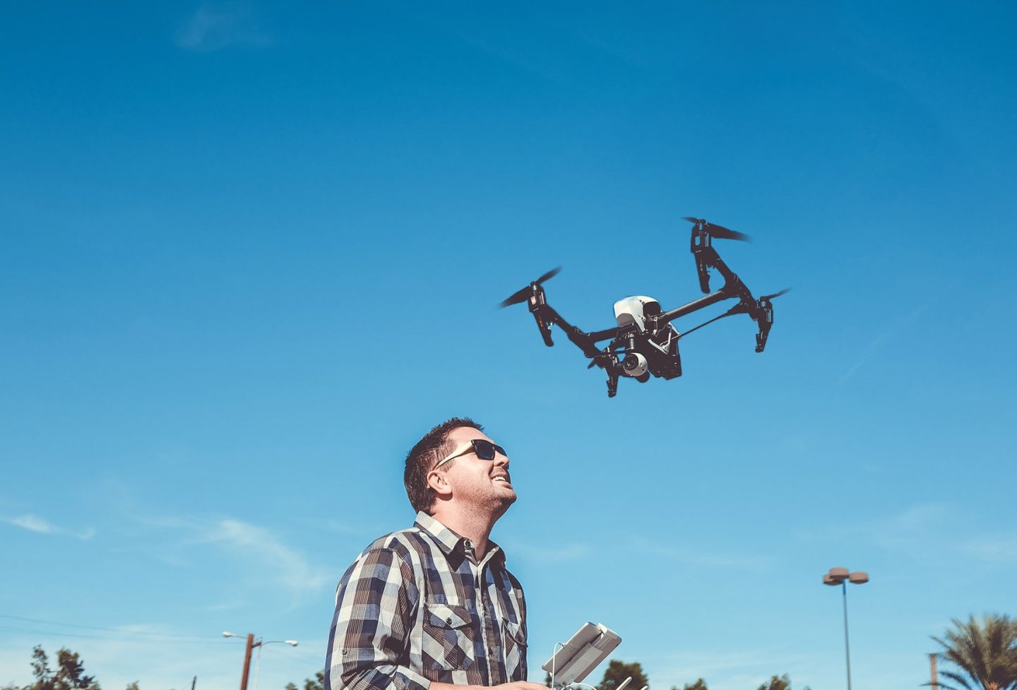 A smiling man in sunglasses using remote control to pilot small drone flying over his head against a blue sky.